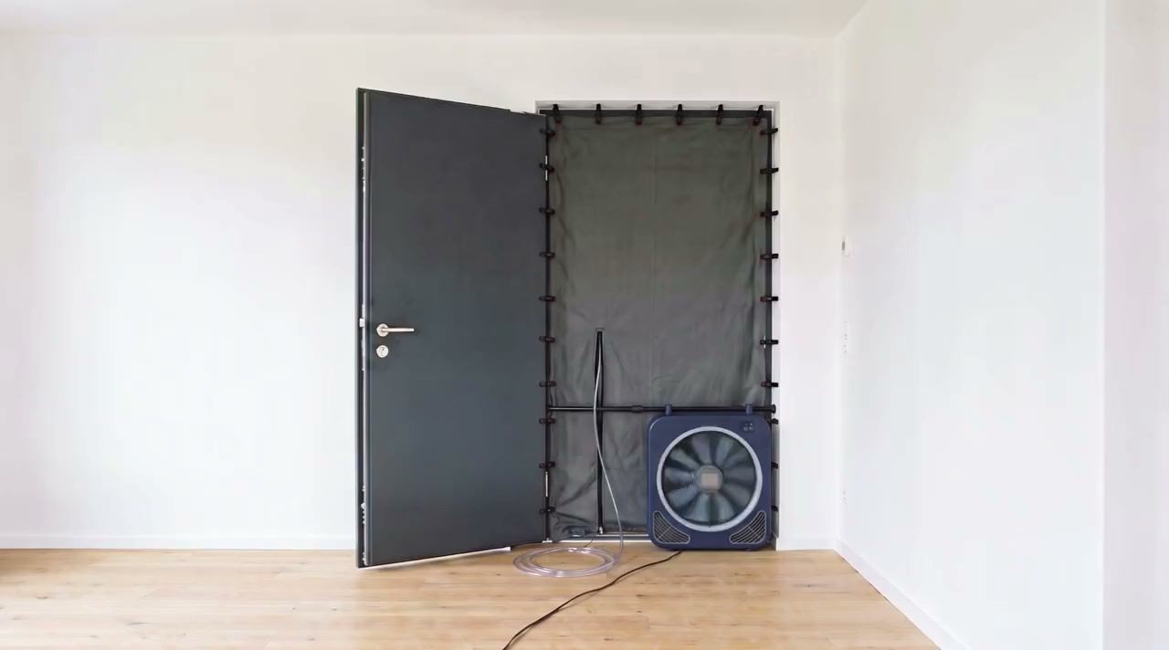 Blower-Door-Messaufbau von innen: Das Messgebläse steht unten seitlich in der geöffneten Haustür, die Restöffnung ist mit grauer Abdichtfolie verschlossen und rundum mit Klammern am Rahmen befestigt; Teleskopstange, Messschläuche und Netzkabel sind sichtbar.